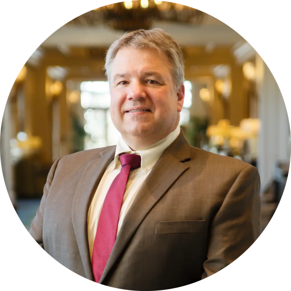 A professional man in a brown suit and red tie smiling indoors.